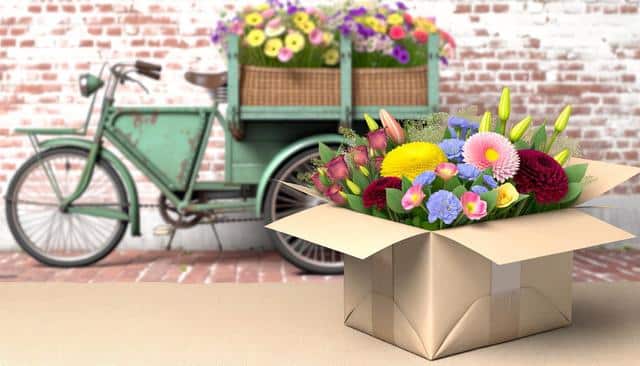 A box of colorful flowers sits on a table in front of a vintage green bicycle with flowers in its basket.