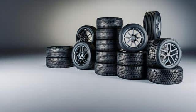 Stacks of black car tires with various tread patterns and alloy rims on a neutral background.