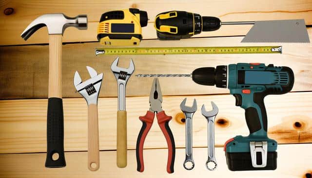 A collection of hand tools and power tools on a wooden surface, including a hammer, wrenches, pliers, saw, drills, and a measuring tape.