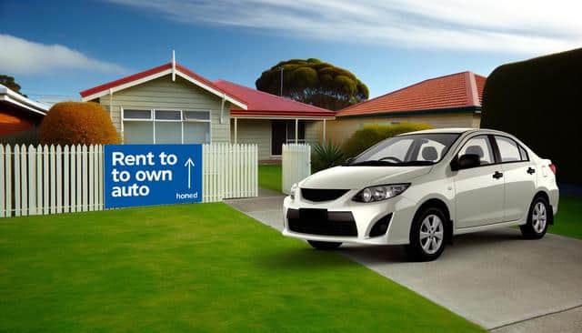 A white car parked on a driveway by a house with a "Rent to own auto" sign on a picket fence.