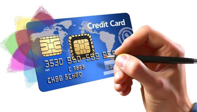 A hand holding a pen, drawing numbers on a blue credit card with colorful abstract shapes in the background.