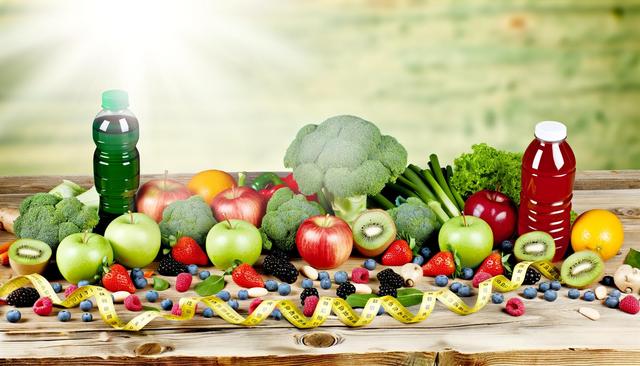 A variety of fruits, vegetables, and drinks on a wooden table with a measuring tape.