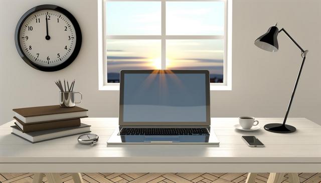 A tidy desk with a laptop, books, clock, lamp, cup, and pencils. Sunlight streams through a window.