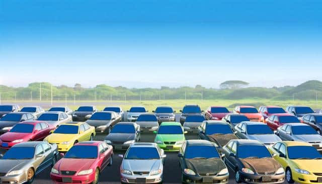 A parking lot filled with various colored cars under a clear blue sky, with trees in the background.