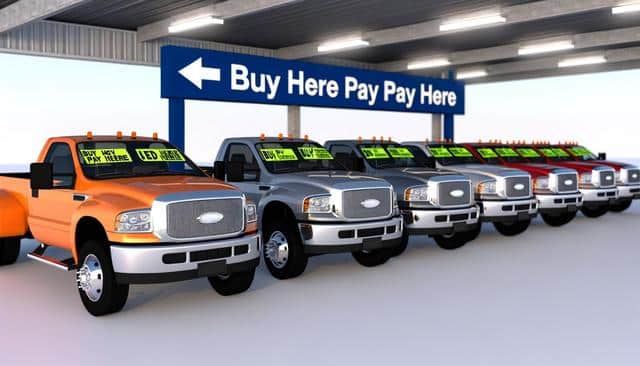 Row of trucks with "Buy Here Pay Here" signs under a carport with overhead lighting.